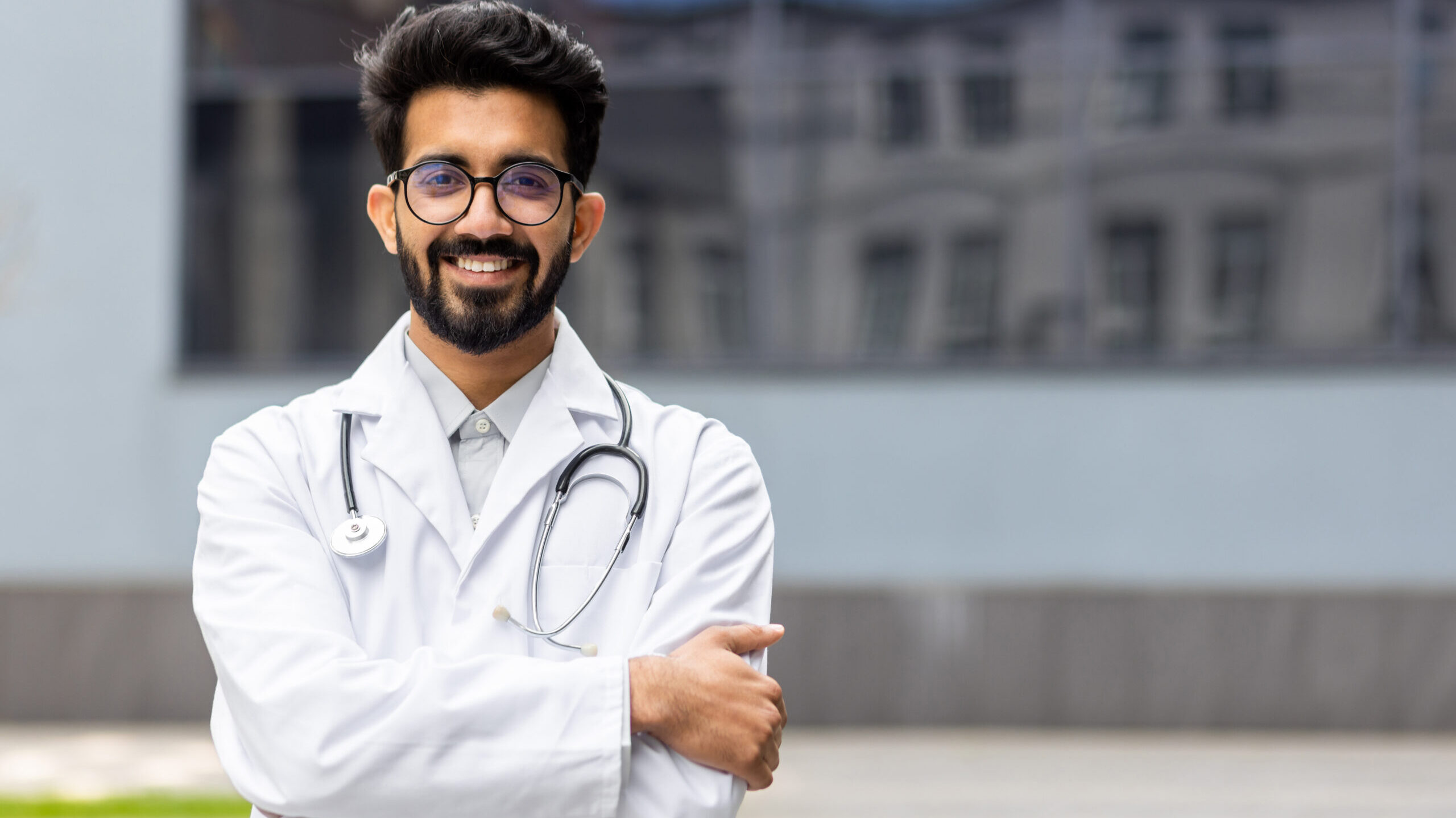 Smiling doctor wearing white coat with stethoscope standing outside hospital